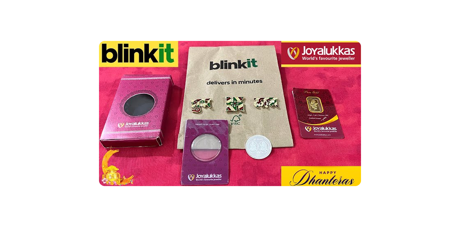 Promotional display of Joyalukkas jewelry packaging on a red cloth, with pink and purple boxes, jewelry cards, a foil bag that says 'blinkit delivers in minutes' in the center, and a coin - Diwali Marketing Ideas